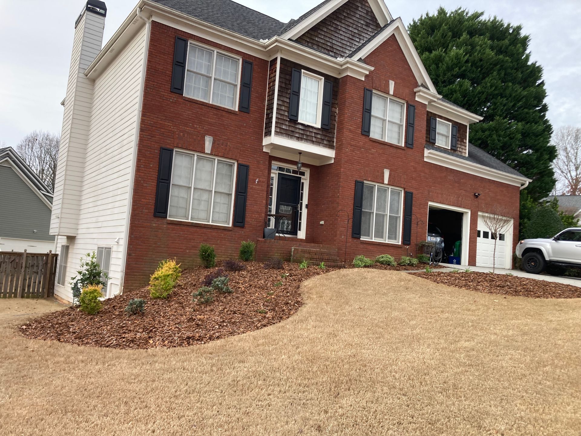Two-story brick house with white siding, black shutters, and a chimney. Landscaped yard with mulch and shrubs.