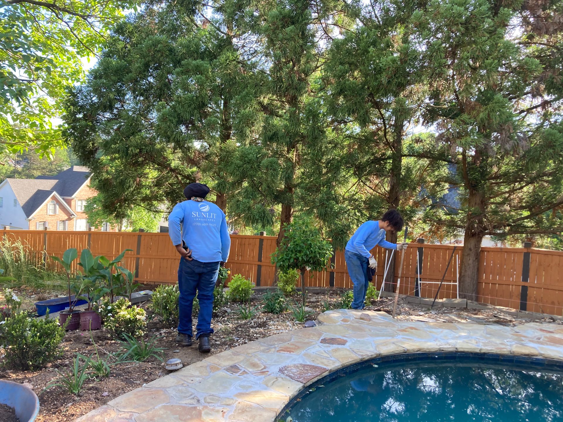 Two people gardening beside a backyard pool and stone patio under trees.