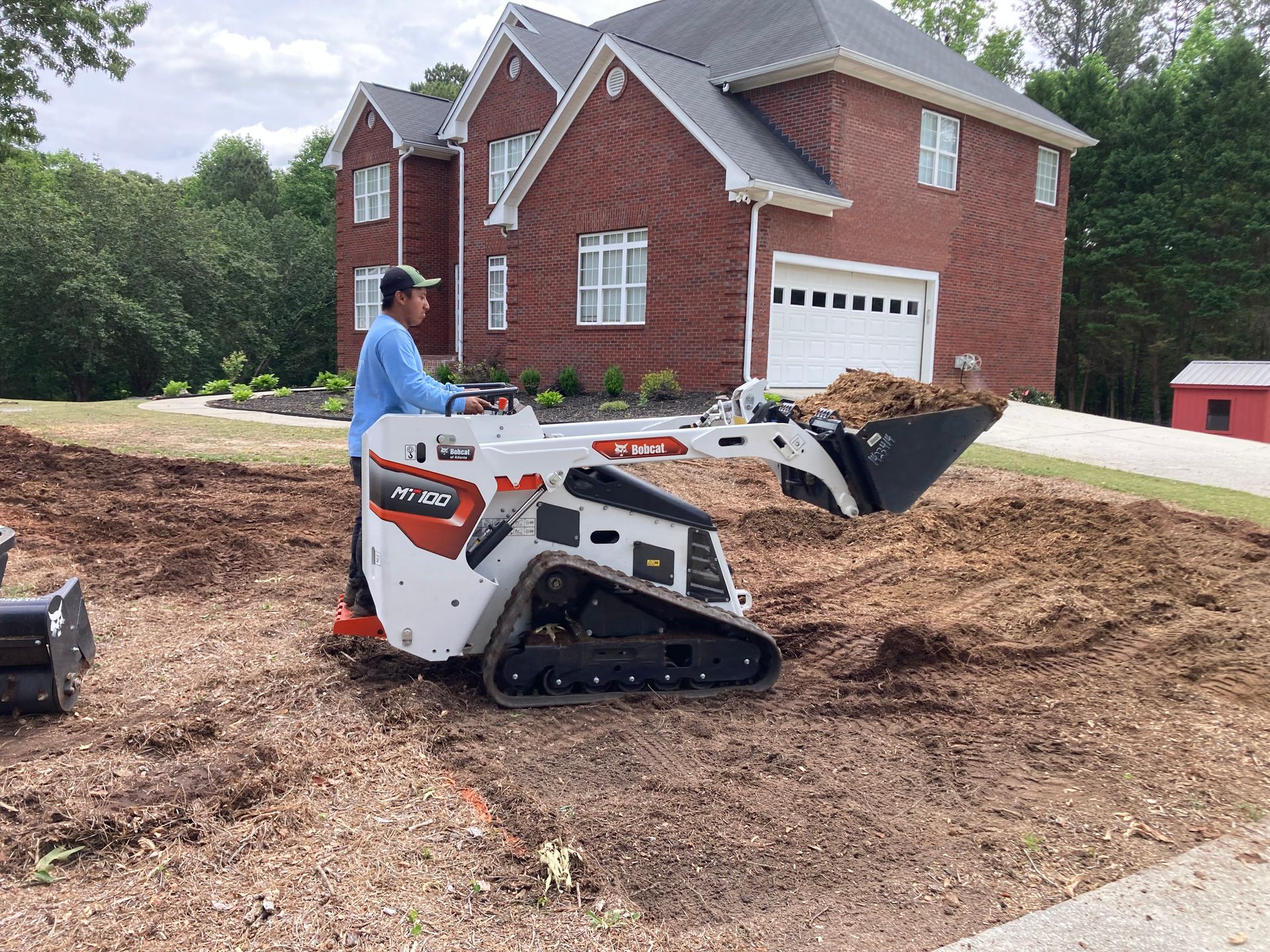 Worker operating a compact track loader on a dirt pile beside a red brick house