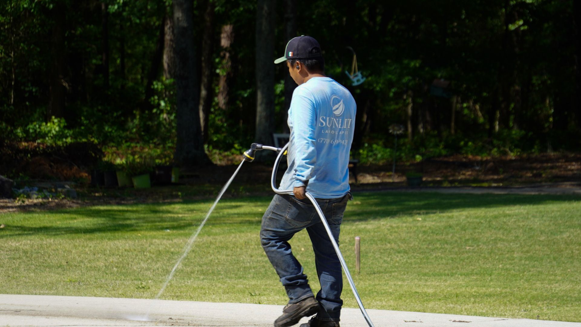 Person in a cap using a hose or pressure washer on a driveway beside a grassy yard.