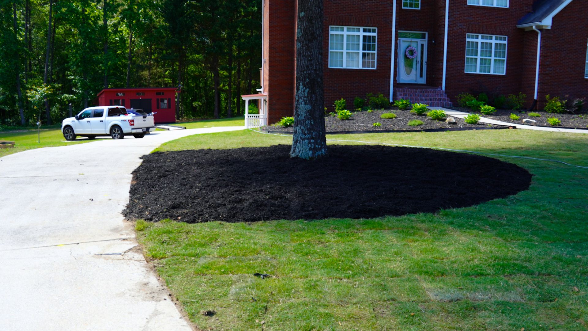 Brick house with a circular mulch bed around a tree beside a curved driveway and parked vehicles