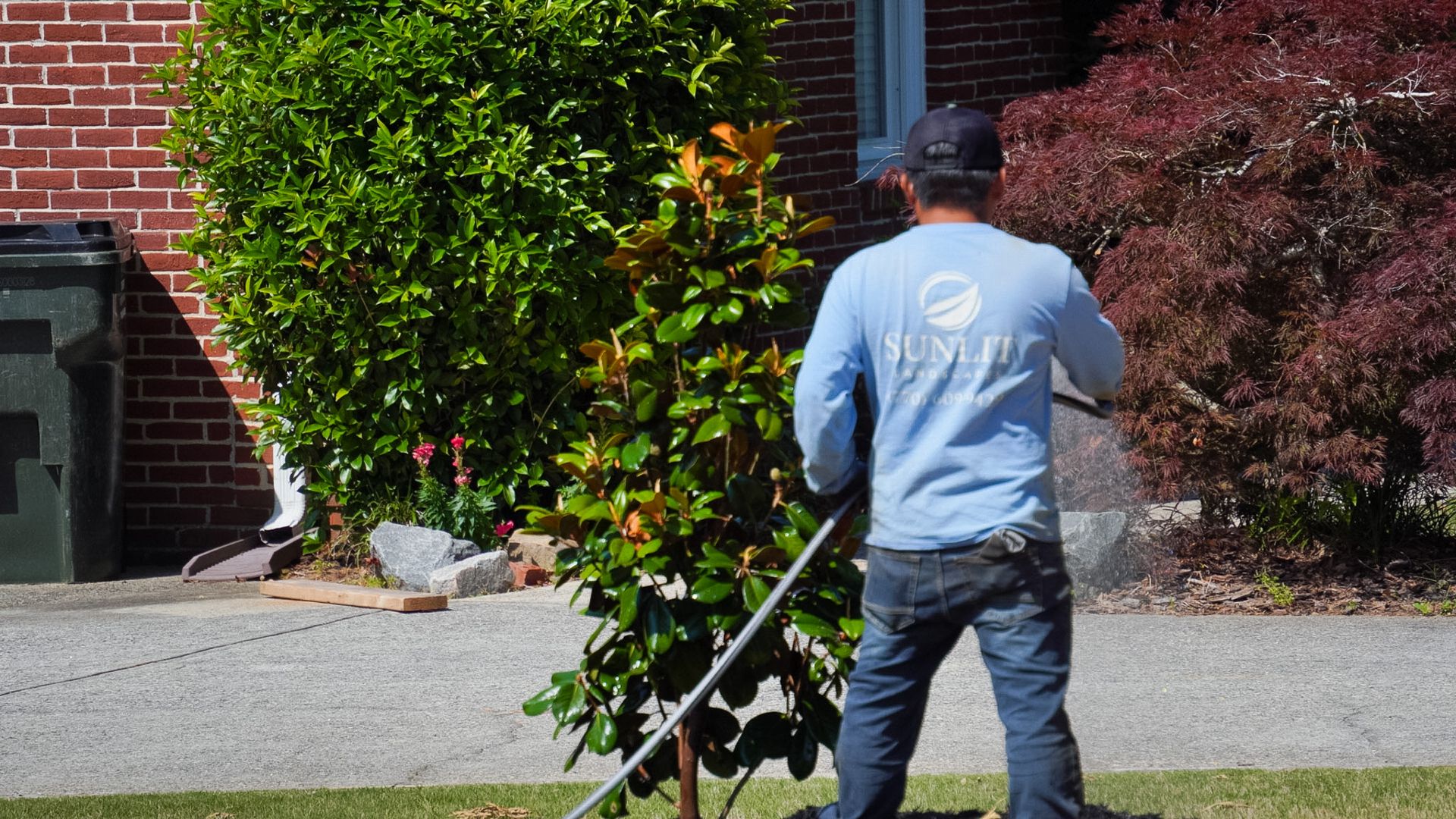 Person in a blue shirt mowing a lawn beside a brick house.