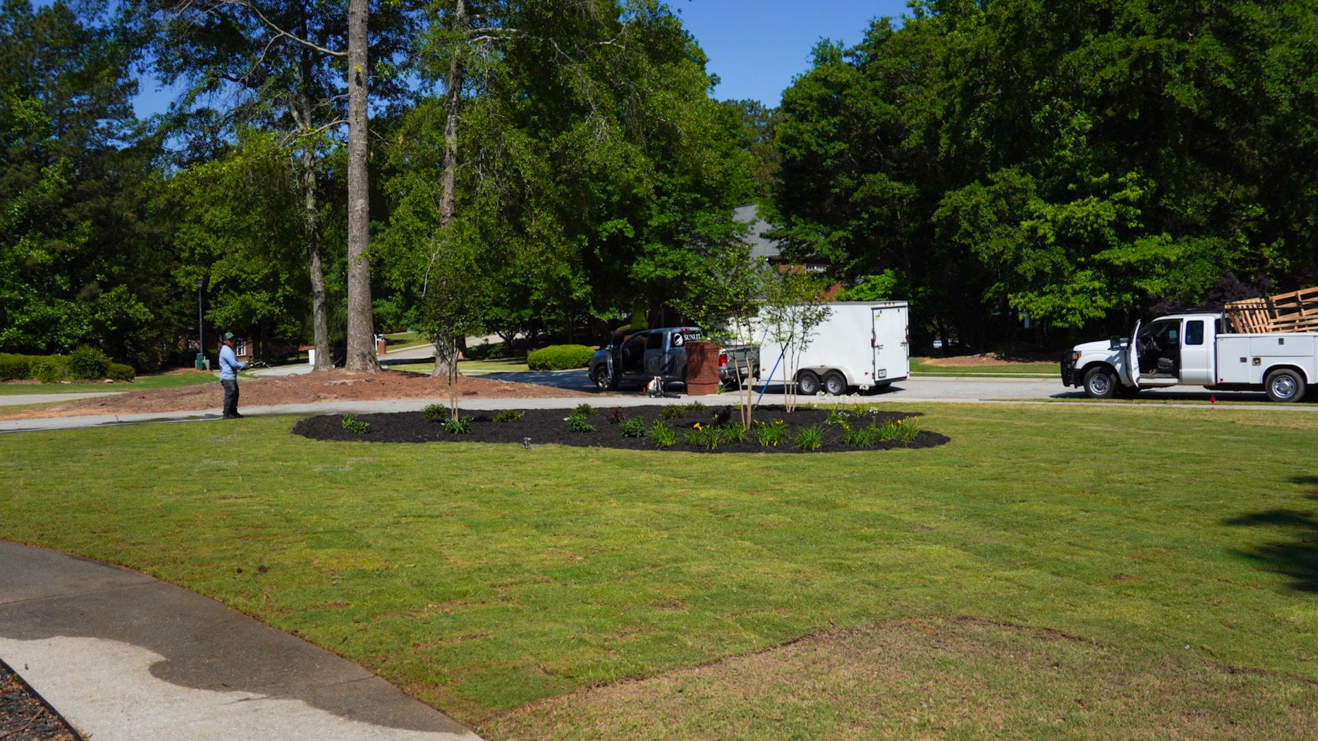 Parkside lawn with trees, a stone flower bed, and white utility trucks parked along a road