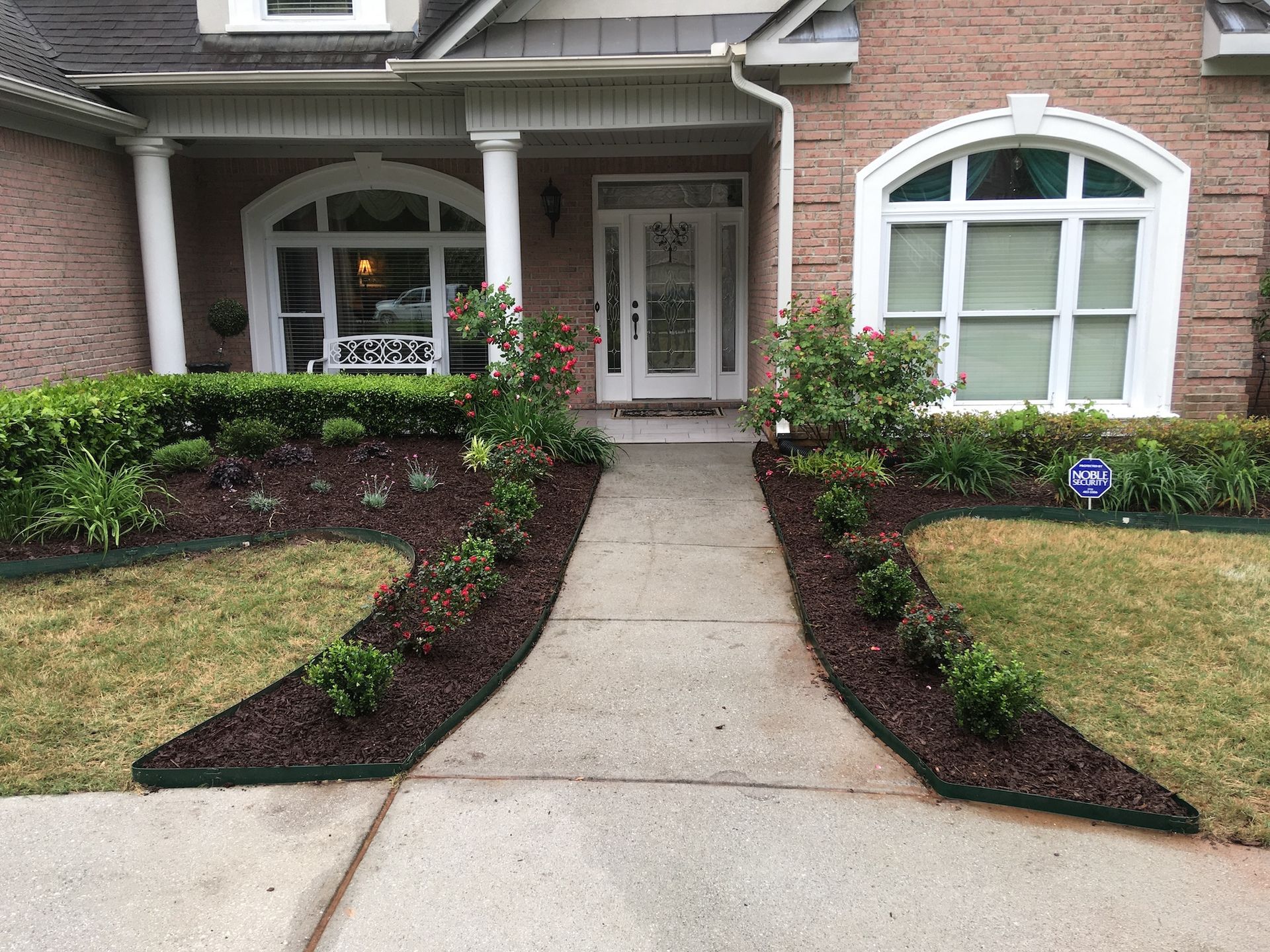 Front yard with walkway flanked by flowerbeds, lush greenery, and a brick house.