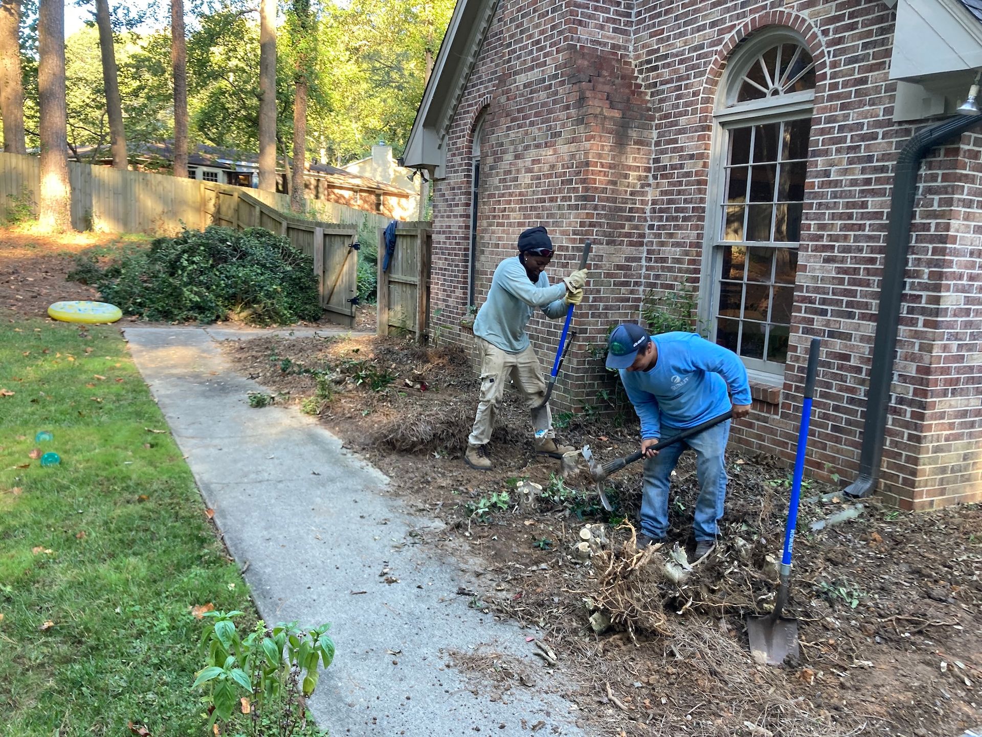 Two people working on landscaping near a brick building, using shovels to remove debris.