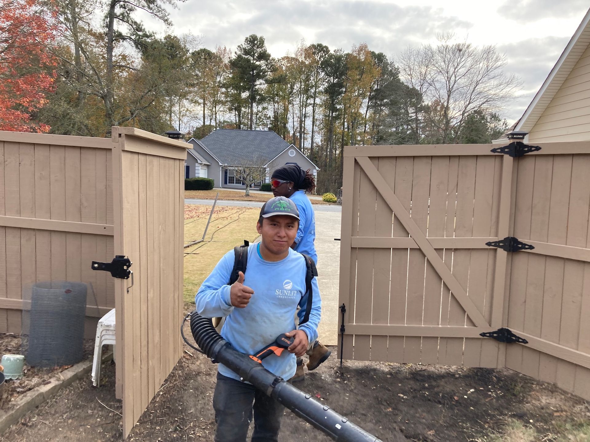 Two workers with leaf blowers inside a wooden fence; one gives a thumbs up.