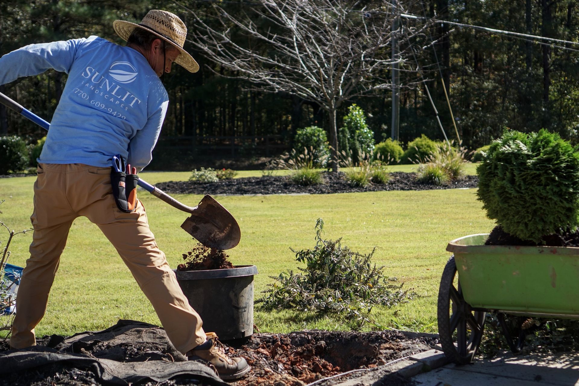 Man in straw hat shoveling dirt from a pot into a hole in a yard; a wheelbarrow with a plant is nearby.