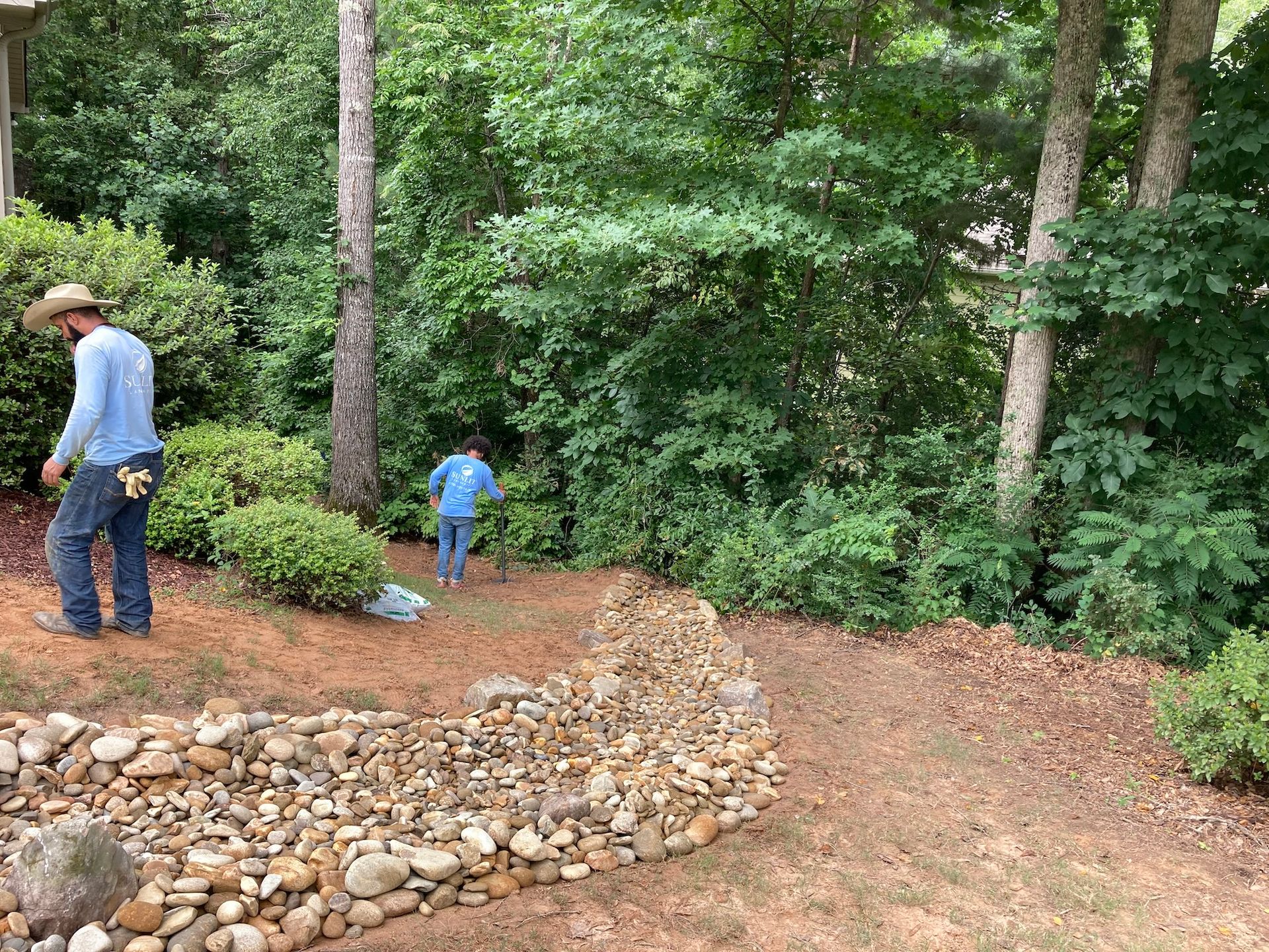 Two workers in a wooded area with a rock border. One is near bushes; the other is further away.