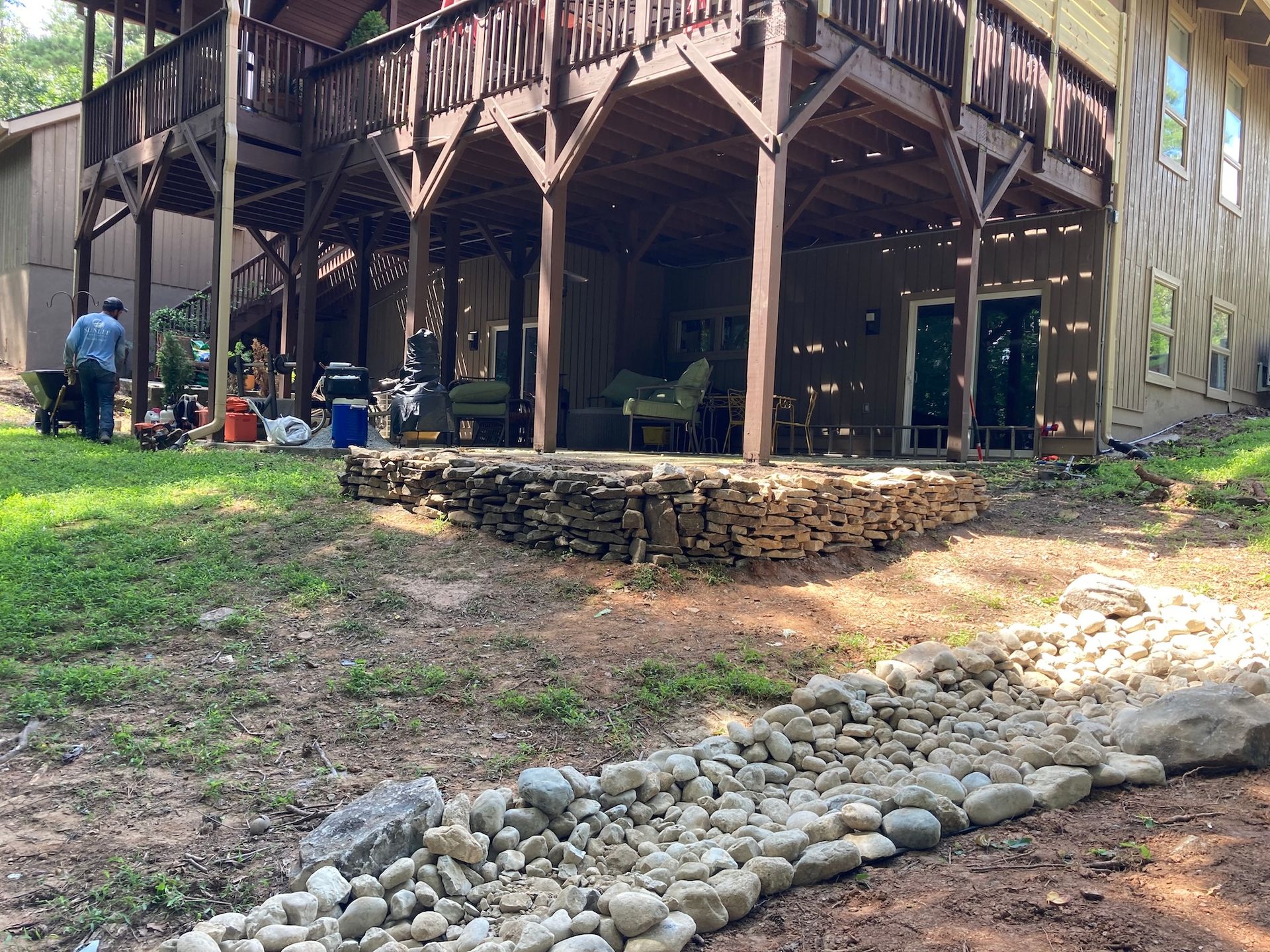 Backyard with a stone-lined garden bed below a raised deck. A man works near the deck.