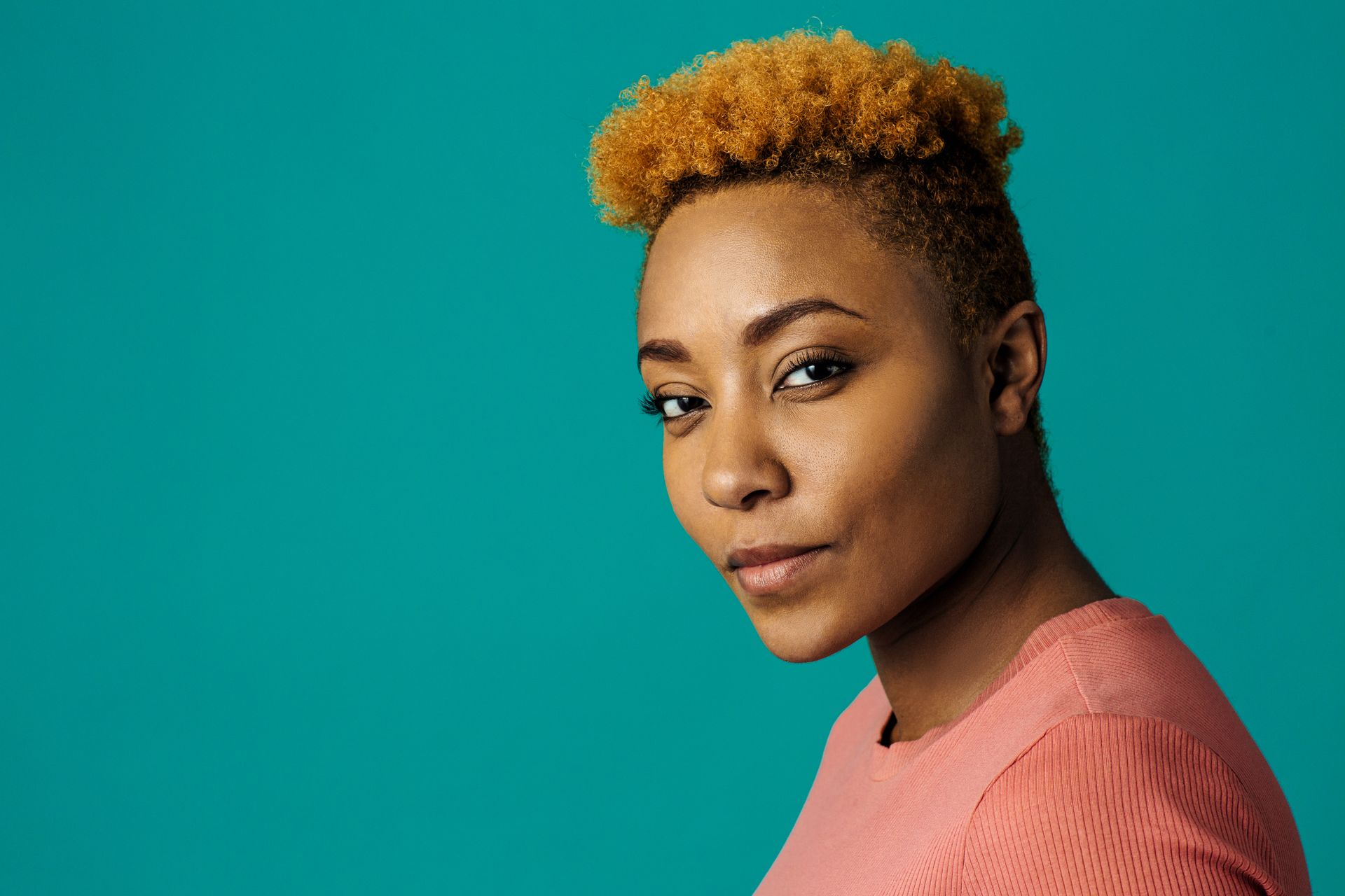 Portrait of a person with short curly hair against a teal background, wearing a coral top, looking at the camera Portrait of a person with short curly hair against a teal background, wearing a coral top, looking at the camera