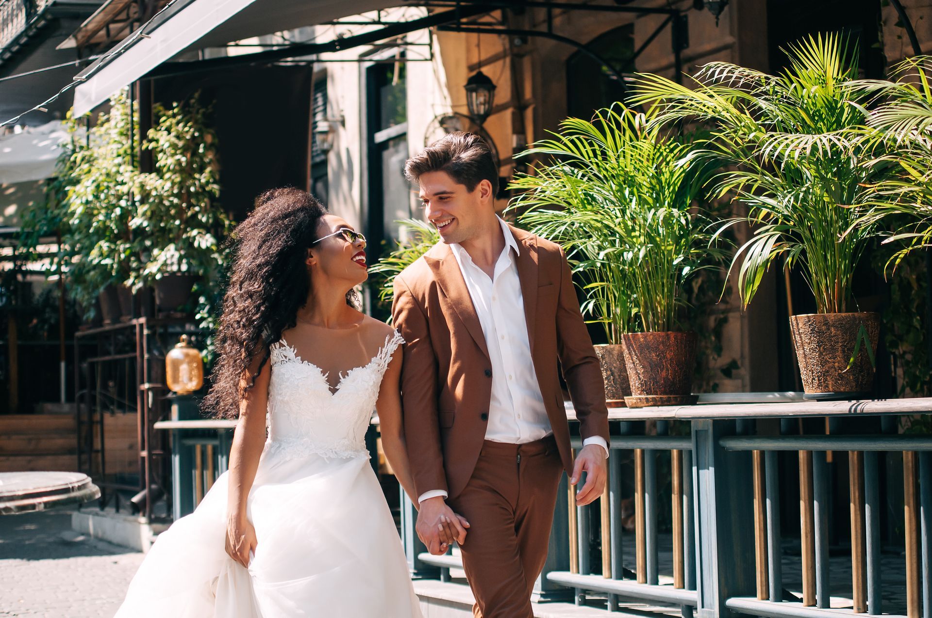 Couple in wedding attire walking hand in hand along a sunny outdoor patio with lush plants