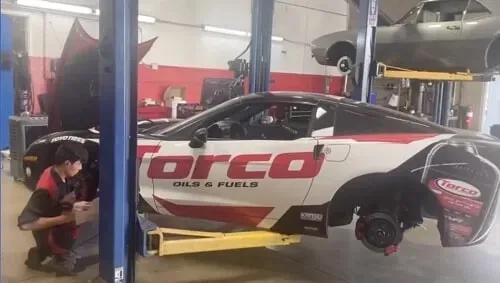 A mechanic works on a white and red Torco-branded car elevated on a lift in an auto shop.