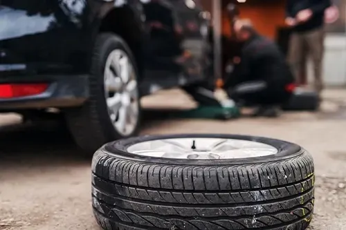 A close-up of a removed car tire in the foreground with a mechanic working on a lifted car in the background.
