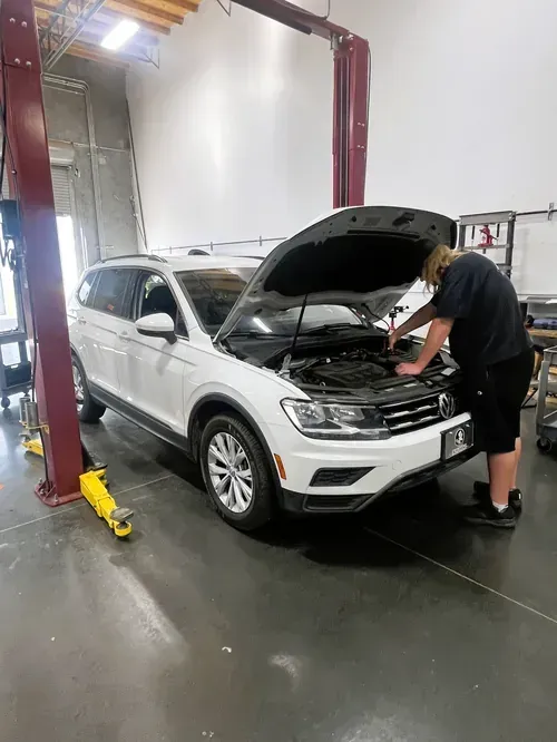 White SUV in an auto shop with hood open while a mechanic inspects the engine on a lift