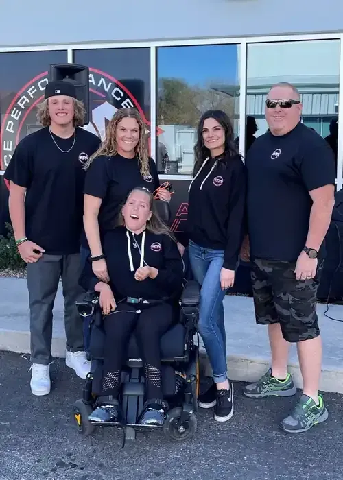 Five people posing outside a storefront, including one seated in a wheelchair, smiling at the camera.