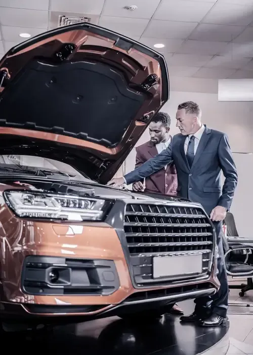 Two men inspect an orange SUV with its hood open in a showroom