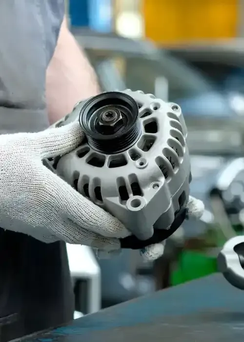Gloved hands holding a car alternator in a workshop, with a blurred vehicle in the background