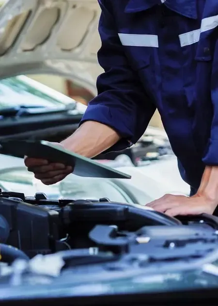 Mechanic in blue coveralls checking a car engine with a tablet, hood open