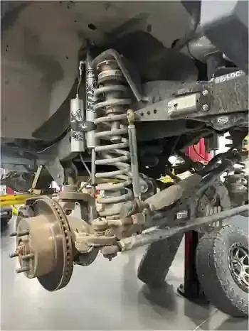 Close-up of a truck’s exposed suspension, coil spring, brake rotor, and wheel assembly in a garage.