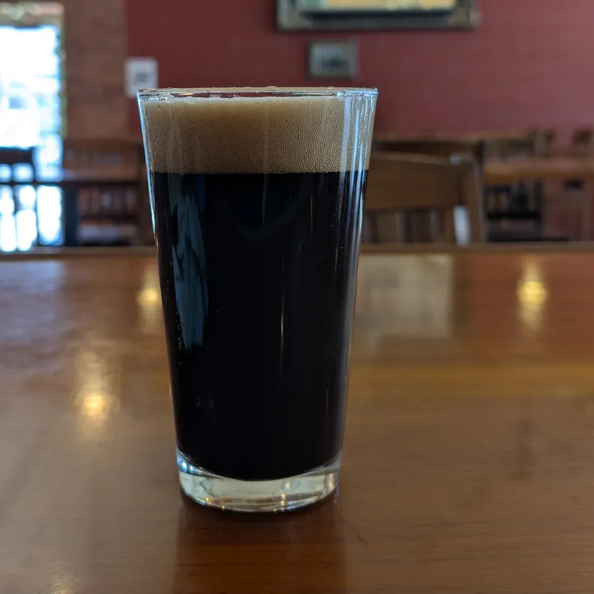 A pint of dark beer with a tan foam head on a wooden table in a pub.