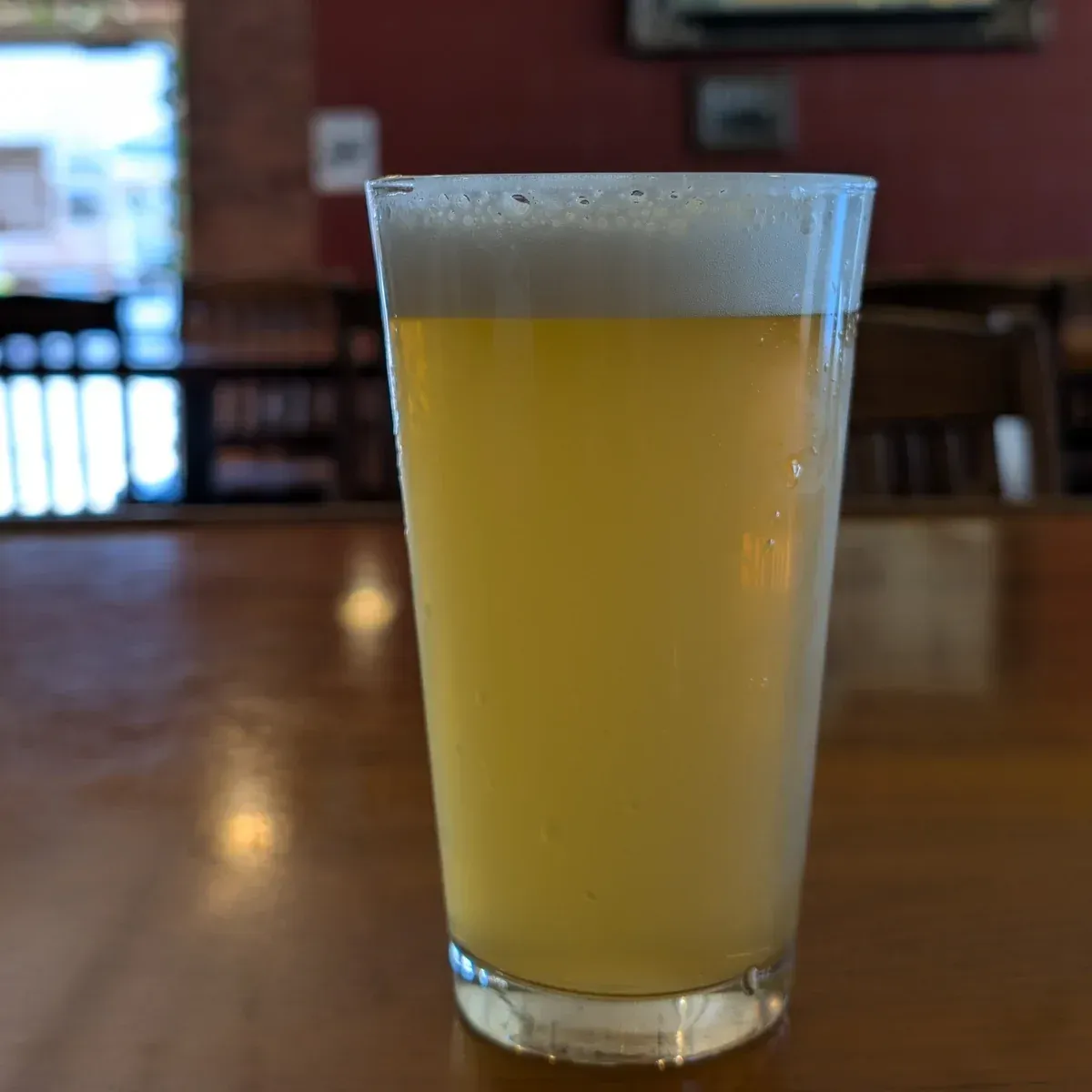 A frothy pale beer in a clear pint glass on a wooden table in a dim bar