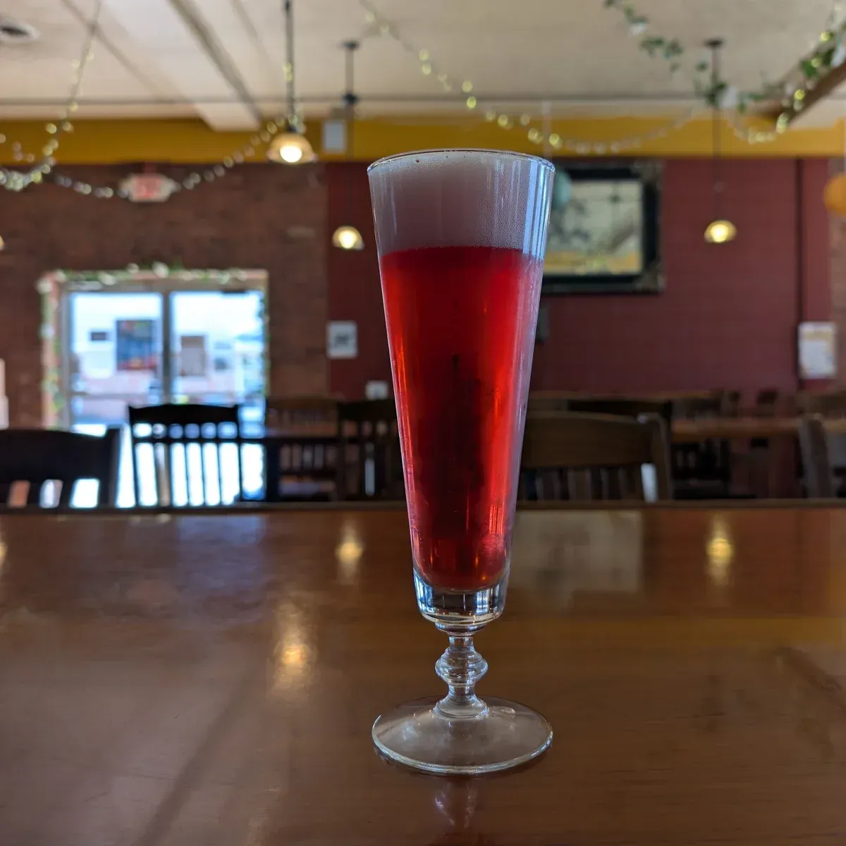Tall red drink in a fluted glass on a wooden table in a restaurant with blurred seating behind.