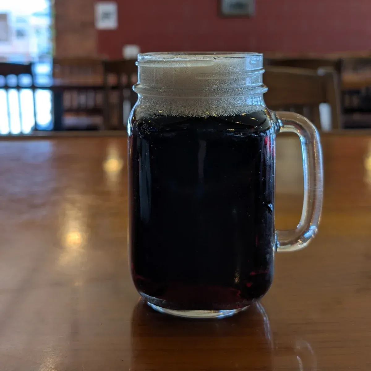 A glass mason jar mug with a dark drink and foam, sitting on a wooden table in a restaurant.