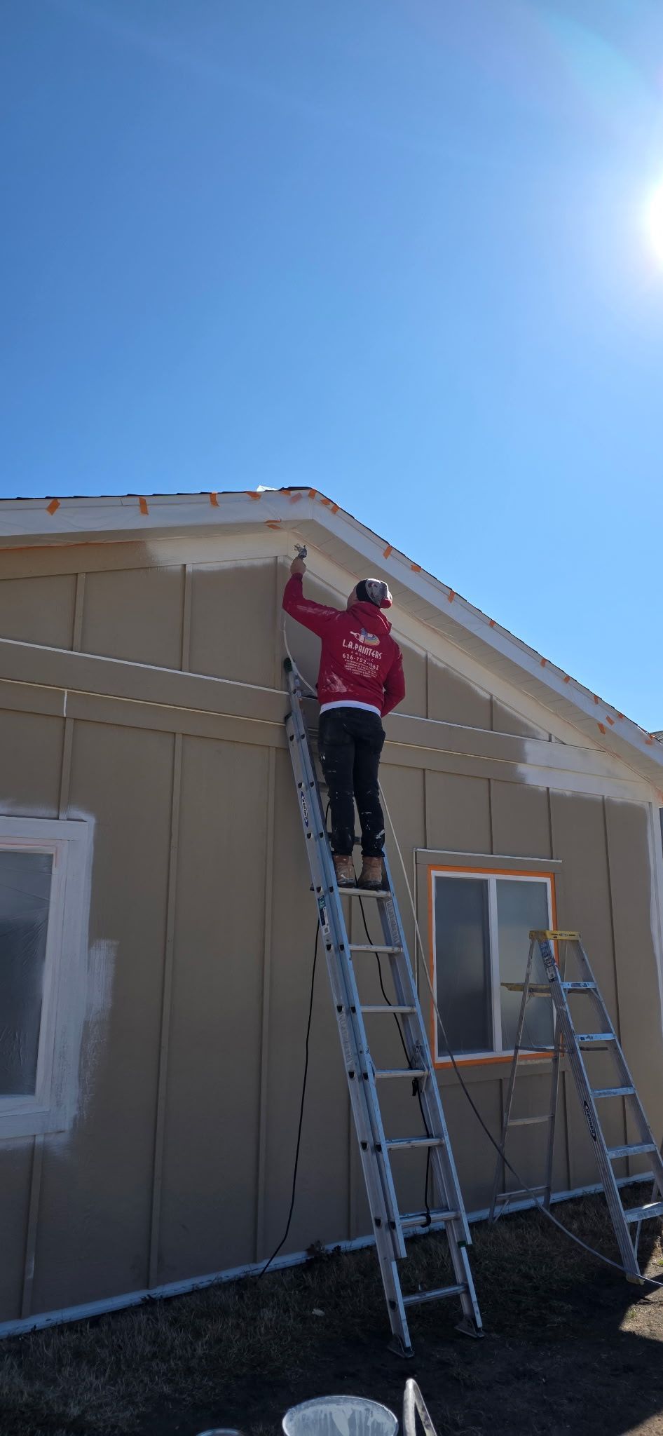 Worker on a ladder painting the exterior of a beige house under a clear blue sky