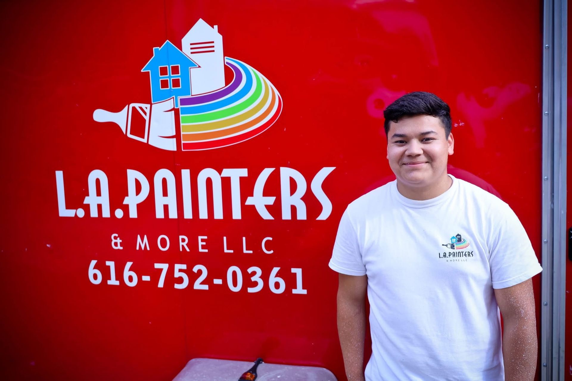 Man standing beside L.A. Painters & More LLC sign on a red wall with phone number 616-752-0361