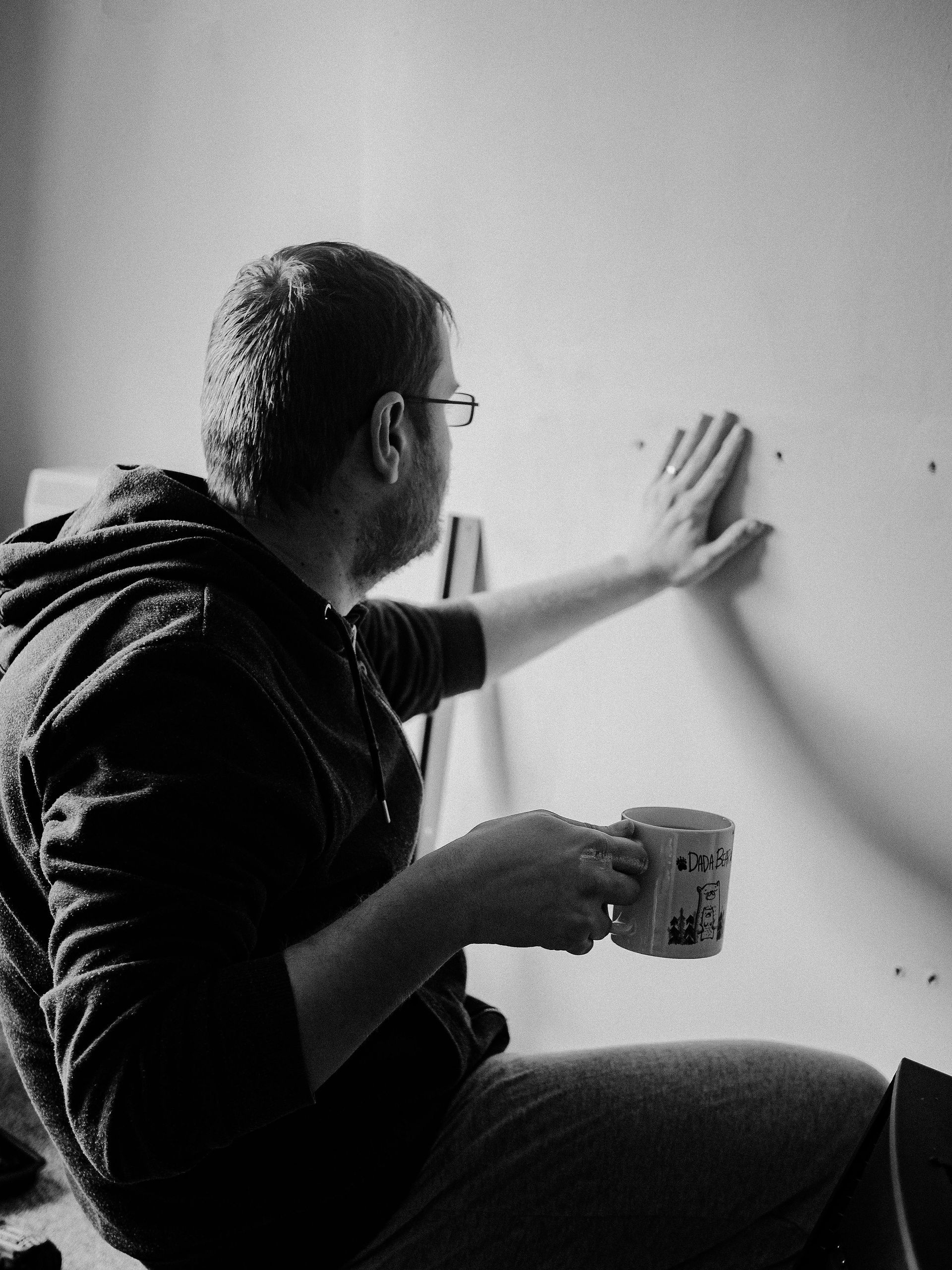 Person kneels by a wall, holding a mug and reaching out to measure or mark the surface.