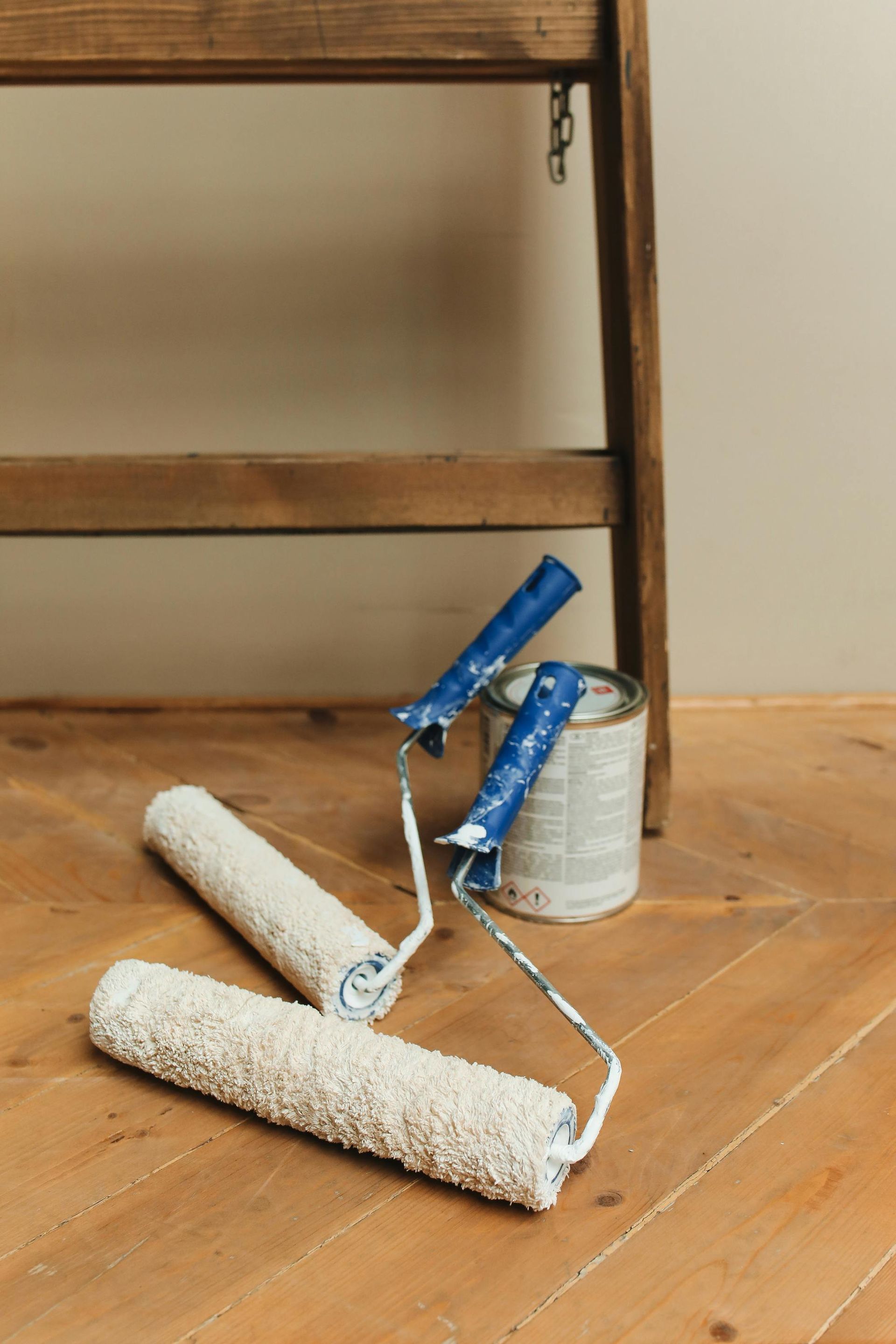 Two paint rollers and a paint can on a wooden floor beside a chair.