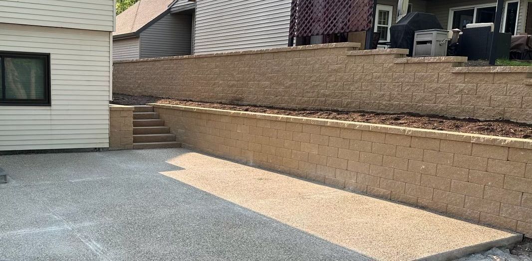 Gravel patio beside a tan retaining wall and house exterior with steps leading up to a raised yard.