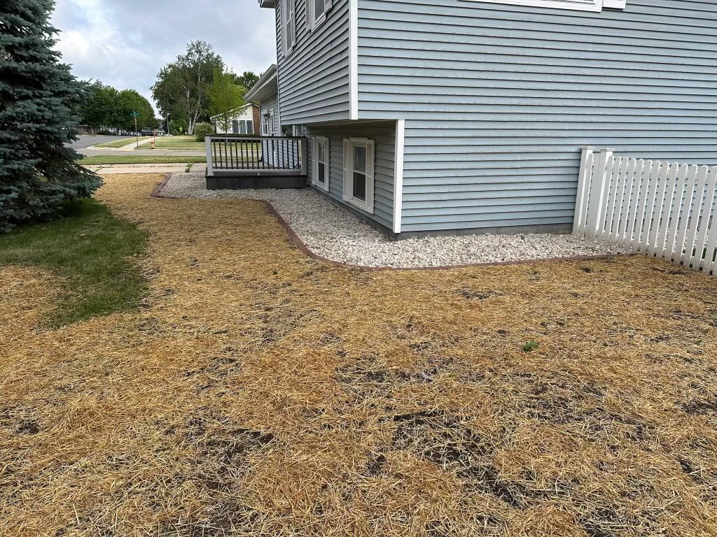 Side of a gray house with a gravel yard, shrubs, and a small deck overlooking a street.