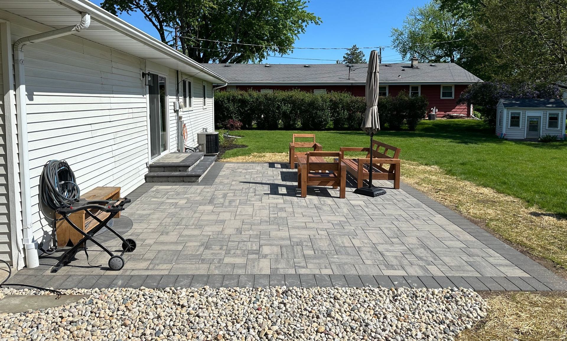Back patio with gray pavers, wooden table and chairs, and a charcoal grill beside a white house