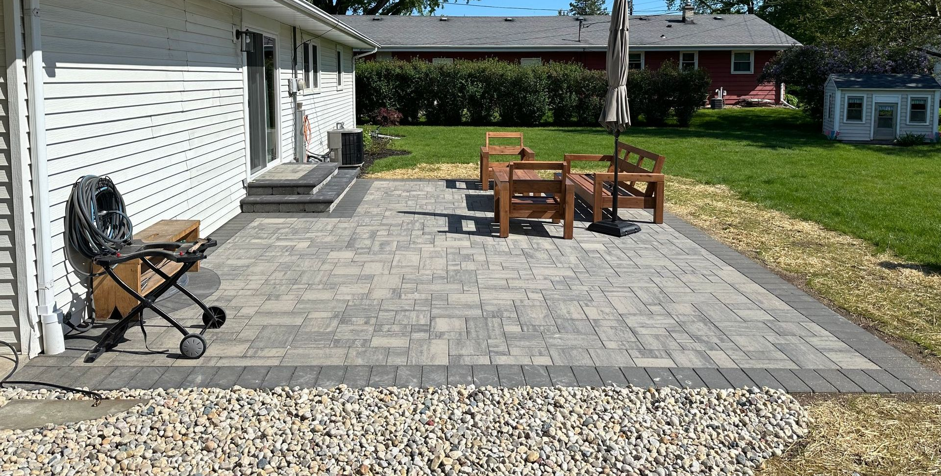 Patio with stone pavers, wooden chairs and table beside a white garage and lawn under a bright sky
