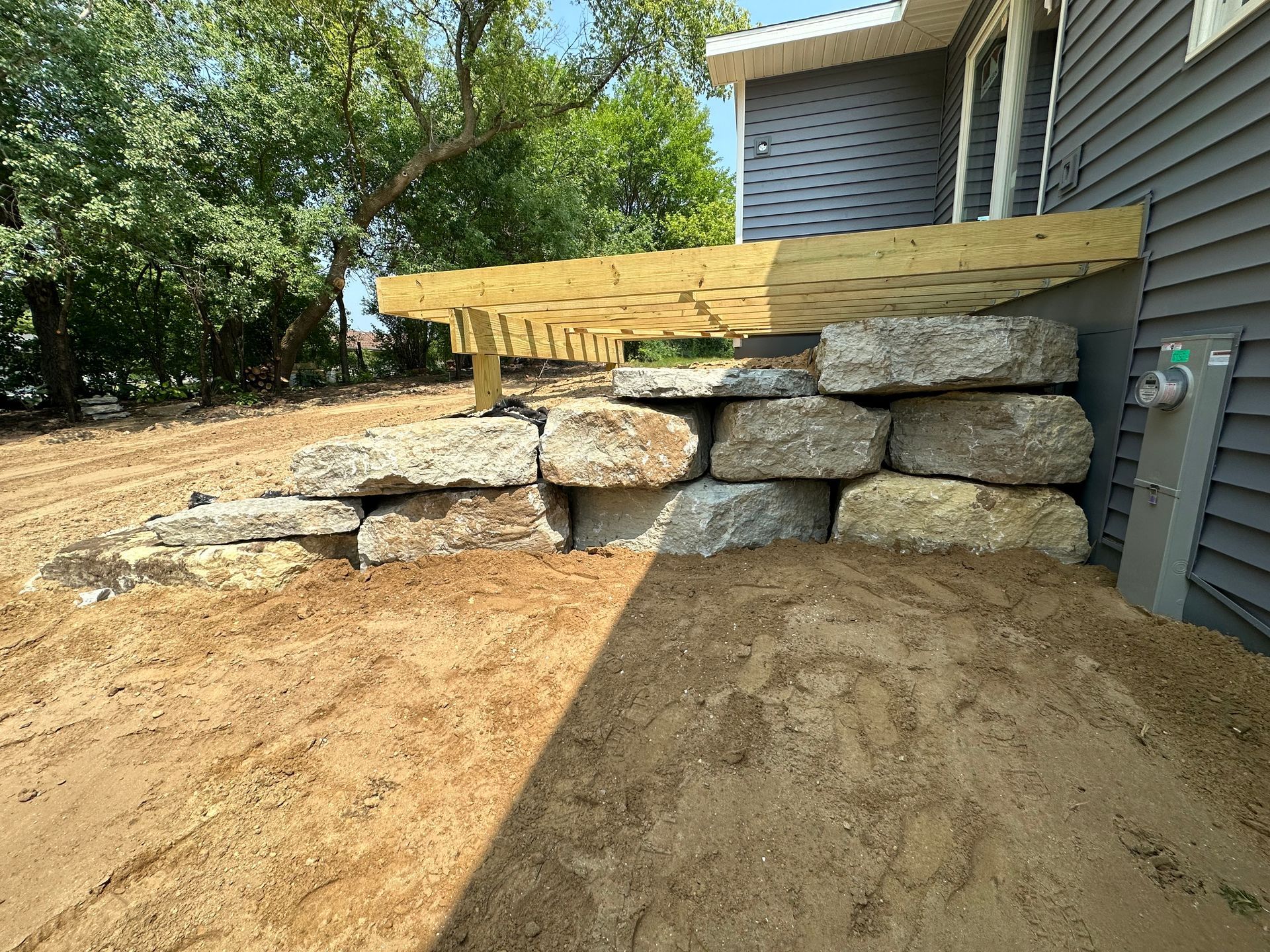 Stone retaining wall beside a house, with a wooden ramp and dirt slope in the foreground