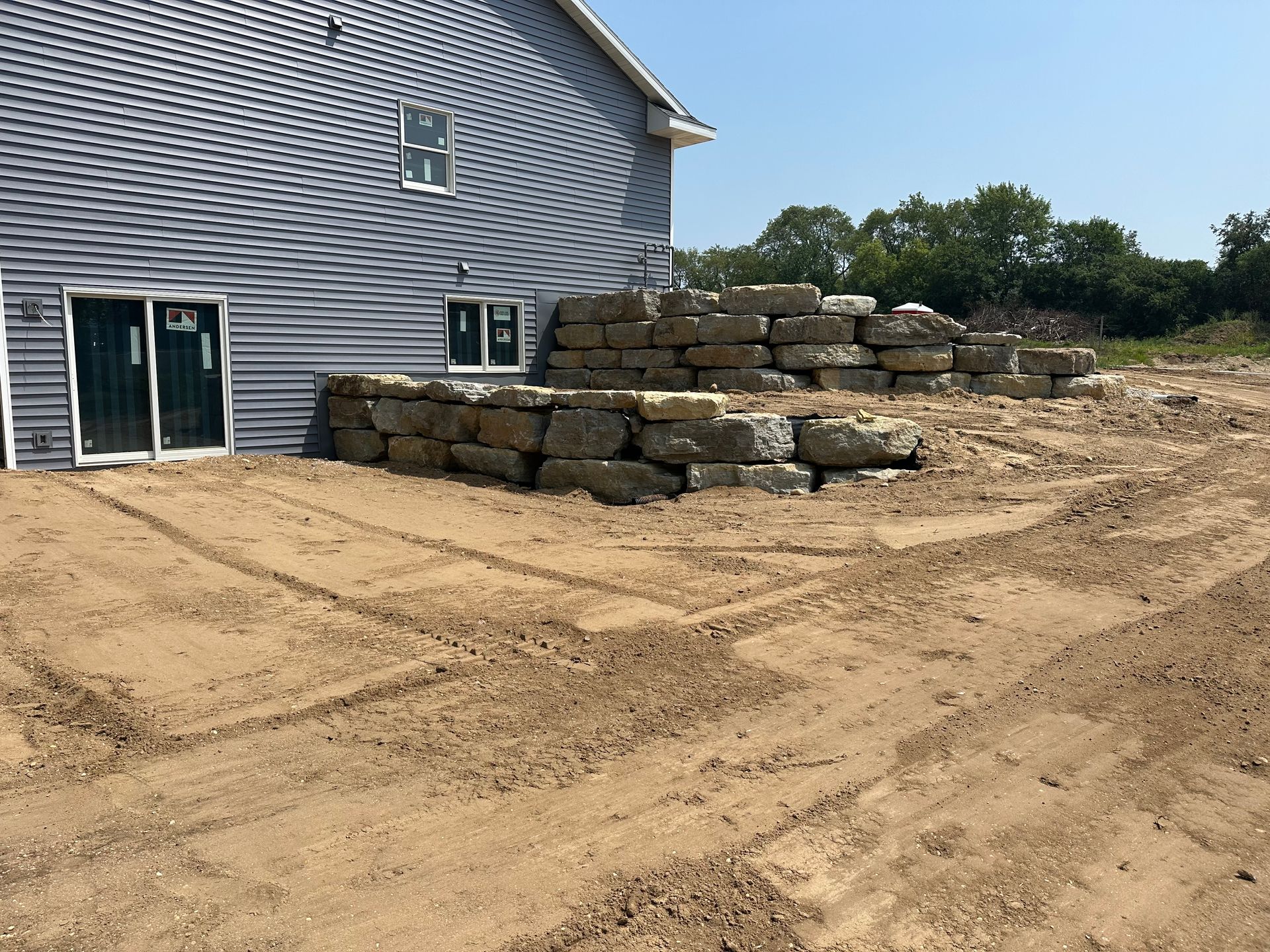 Large stacked stone blocks beside a gray house on a sandy construction site