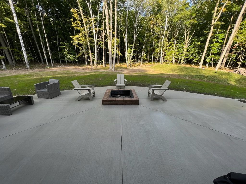 Patio with a square fire pit, lounge chairs, and a wooded backyard beyond