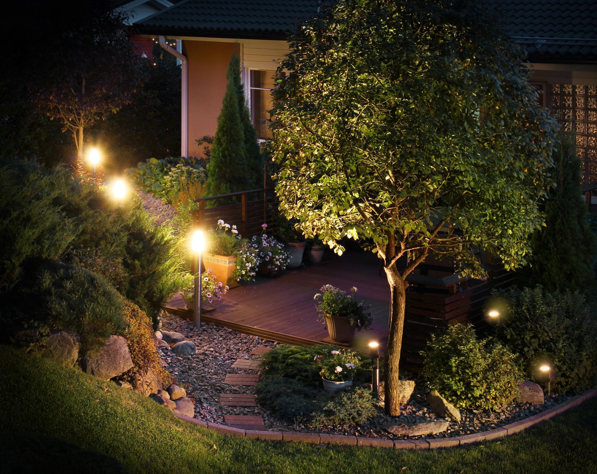Lit garden path and porch beside a house at night, with glowing lamps and a tree-lit courtyard