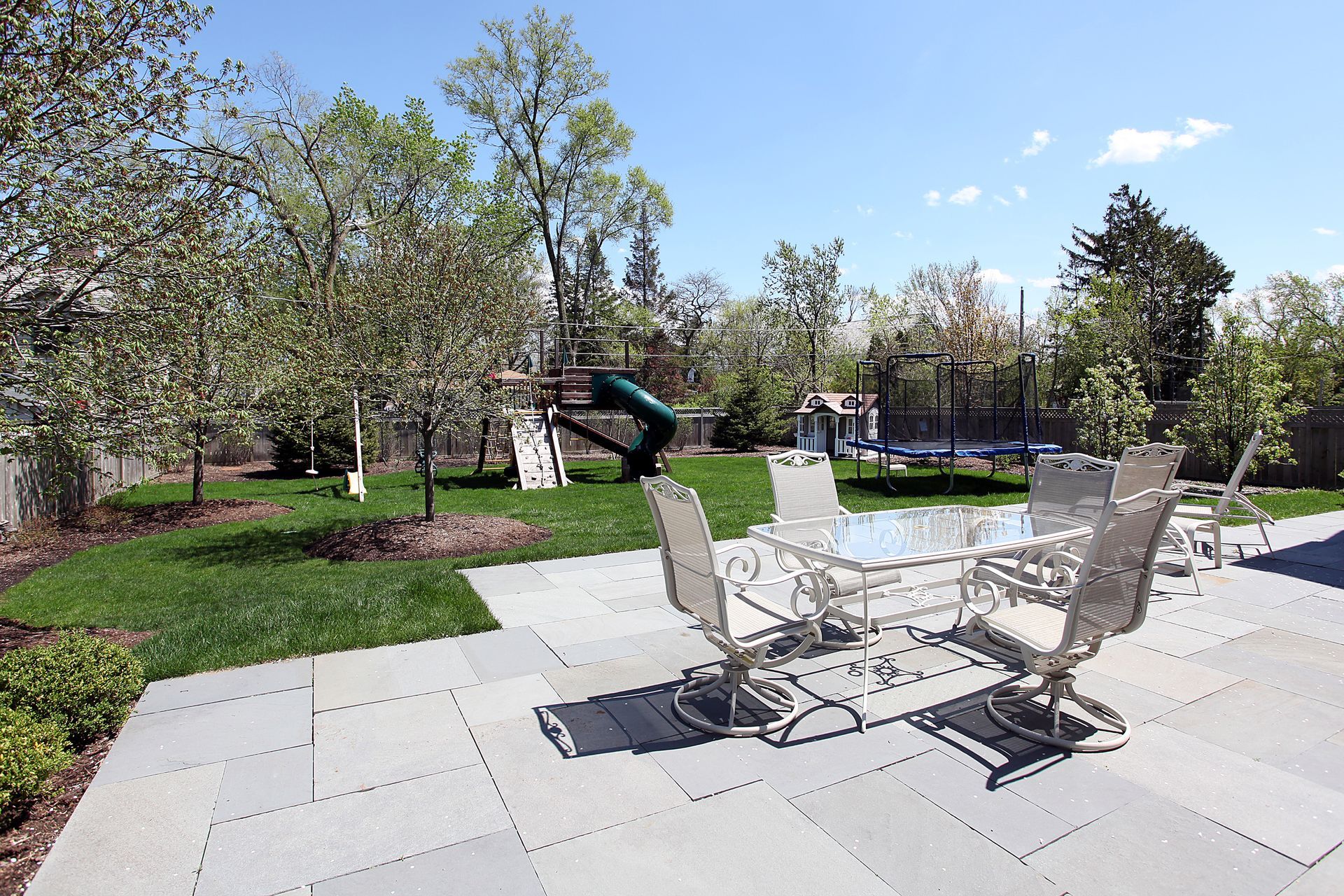 Patio with white chairs and table overlooking a green park with trees and a playground