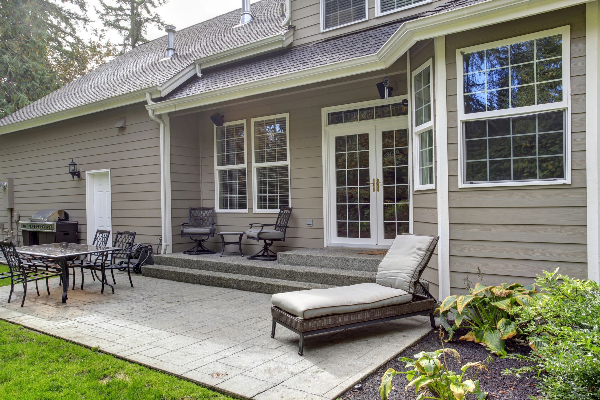 Patio with outdoor seating beside a beige house and glass doors, overlooking a green lawn