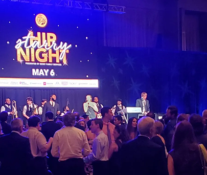 Concert stage with performers and audience under blue-purple lights, “Hire Night” sign and May 6 backdrop.