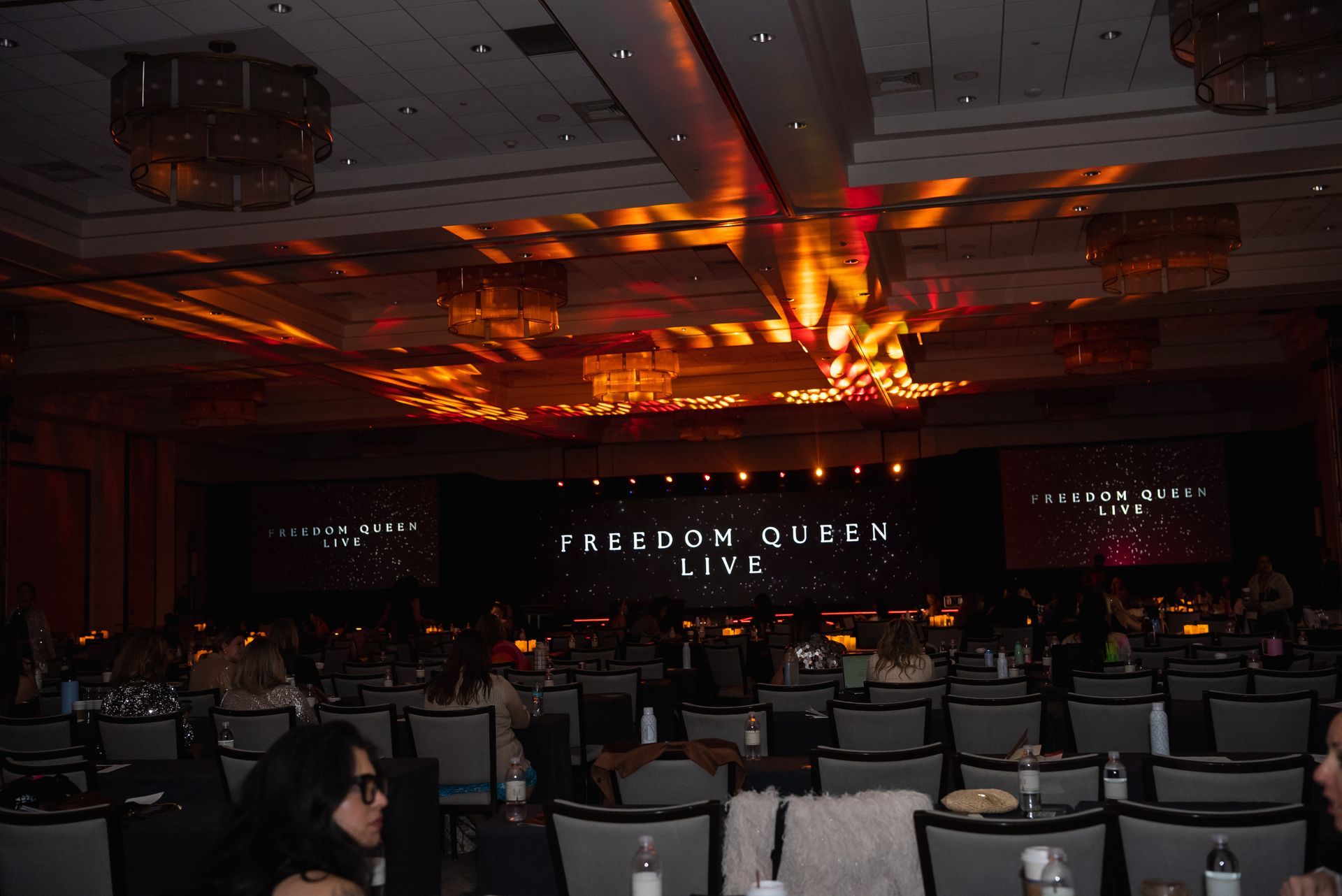 Conference room with rows of chairs facing a stage lit by orange ceiling lights and a large screen.