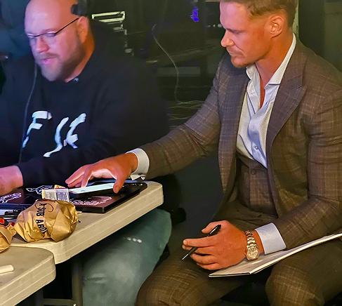 Two men seated at a table, one reaching for a remote beside snacks in a dim room.