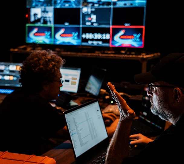 Two people in a dim control room reviewing monitors and a laptop with charts and video feeds
