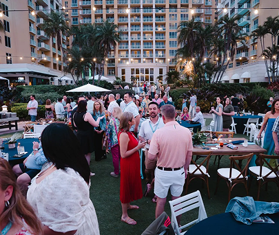 Crowded outdoor poolside cocktail reception at a hotel with guests mingling at tables and standing near the pool.