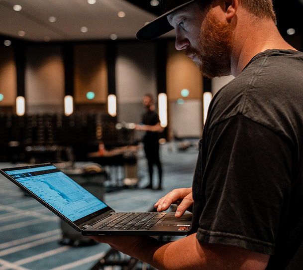 Person using a laptop in a conference hall, with rows of chairs and warm overhead lights in the background