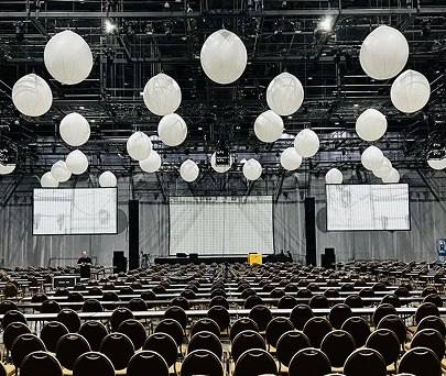 Empty auditorium with rows of chairs, large hanging white spheres, and two projection screens on a stage