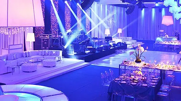 Blue-lit banquet hall with lounge seating, stage lights, and round dinner tables set for an event