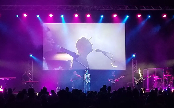 Concert stage with singer silhouette on a bright screen, colorful purple-blue lights, and crowd below